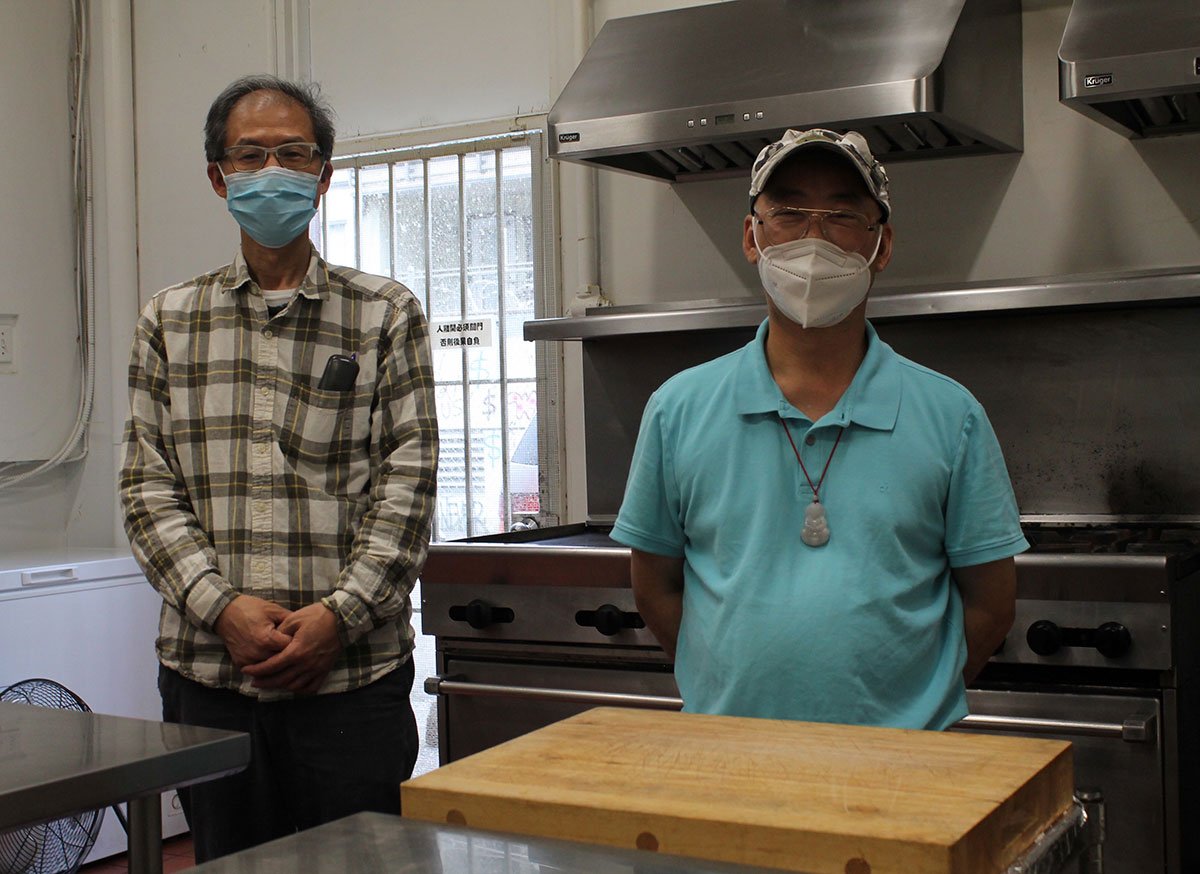 Two Chinese men stand in a commissary kitchen in front of a stainless hood range and behind two clean countertops. Sean Ouyang, left, is in a plaid shirt and medical mask. Floyd Wong, right, is in a turquoise golf shirt, cap and medical mask. 