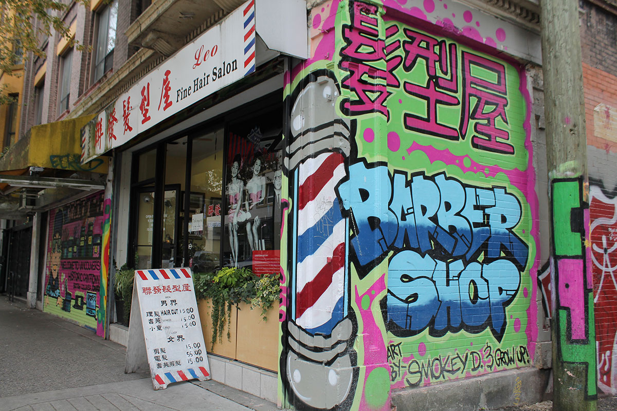 A graffiti mural by a Downtown Eastside street artist Smokey Devil of a blue and red barber shop poll is next to graffiti lettering for “Barber shop” in blue with Cantonese characters in pink above it. 