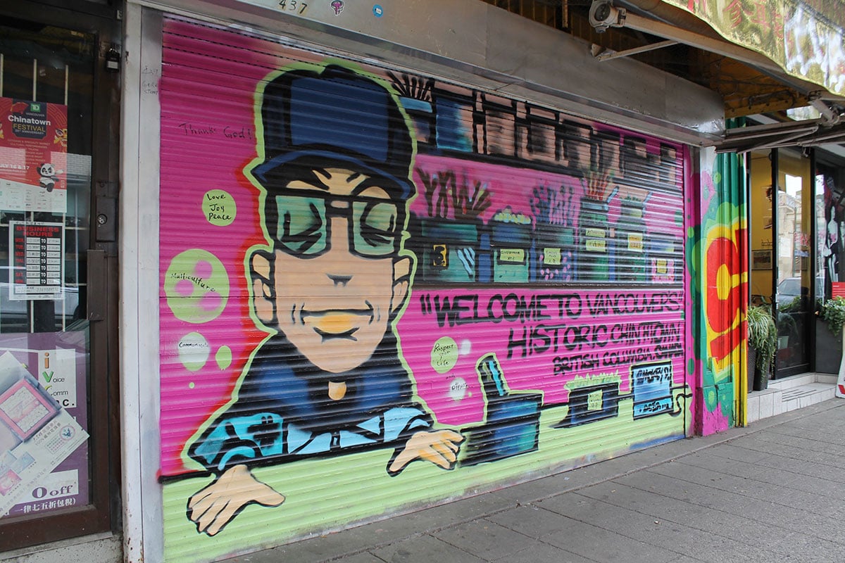 A stylized mural of a Chinatown herbalist in blue clothing and green glasses situates him next to an apothecary shelf. The writing in black text against a pink wall reads: “Welcome to Vancouver’s historic Chinatown, British Columbia, Canada."
