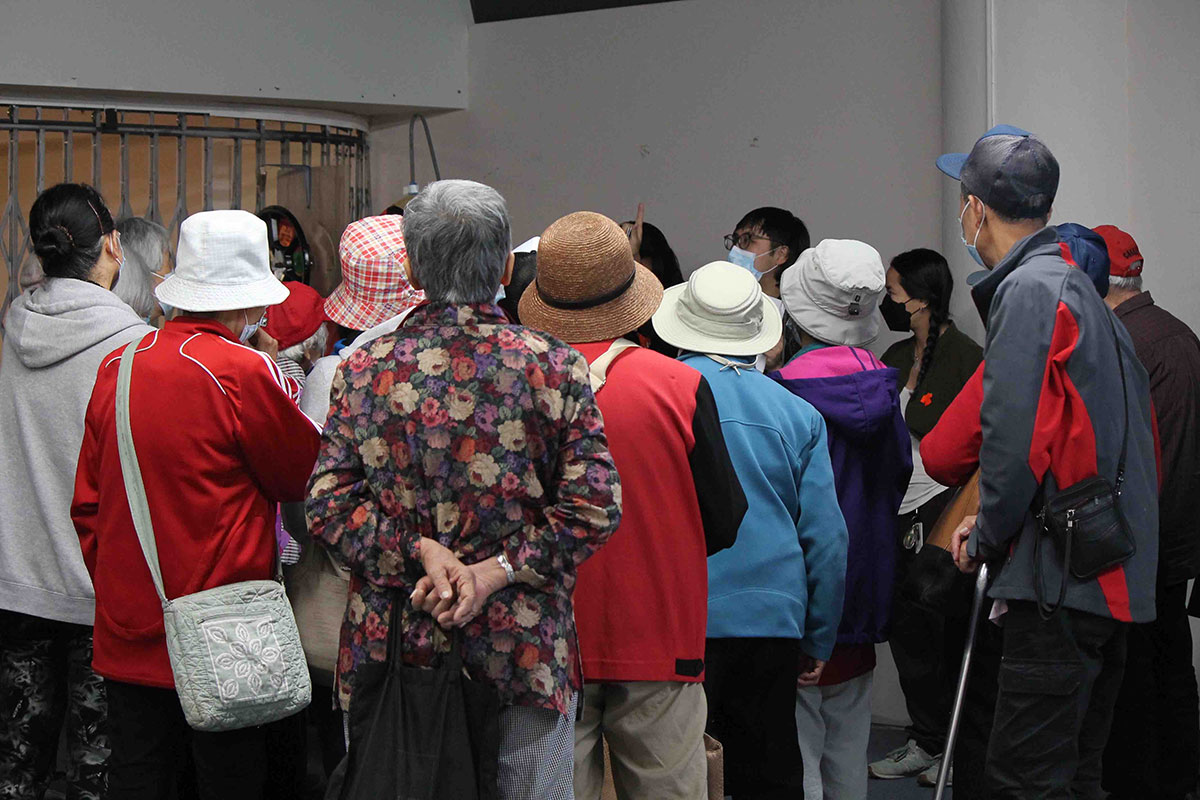 A group of Chinese seniors gathers in a crowd. They are looking at a young Chinese person in a medical mask and glasses, pointing up. The seniors’ backs are turned to the camera. Many are wearing bucket hats and colourful jackets.