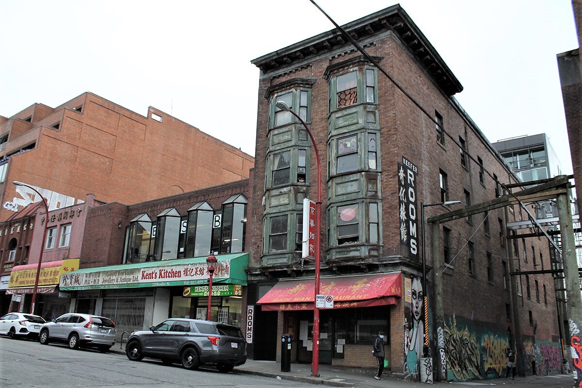 A Chinatown streetfront depicts the Keefer Rooms, a four-storey single-room occupancy hotel with dark green window trim and brown brick. Below it is the Gain Wah Restaurant with a red awning. Next door is a single-storey restaurant called Kent’s Kitchen with a red and green sign.