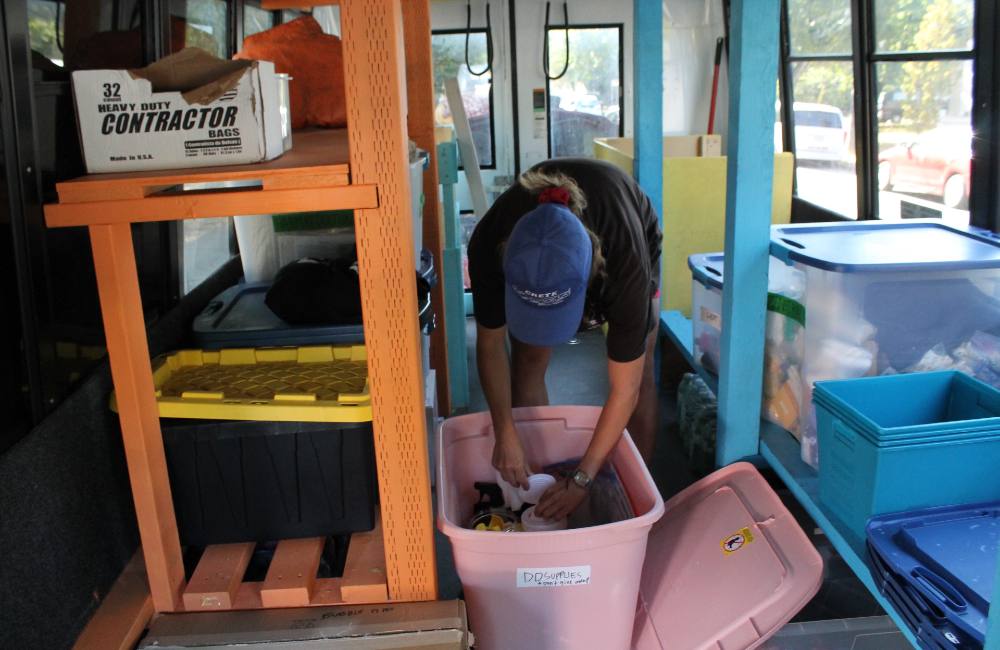 Three stacked photos show the Distro Disco donation hub in action. Top: a spraypainted sign leaning against a grey folding table reads “Distro Disco Mobile Store” in red, green and blue lettering. Middle: a view of the Distro Disco bus, a repurposed handyDart-style shuttle bus with a large waterproof poster hung across its passenger windows in yellow, orange and green colourways. The sign explains what Distro Disco is about. Bottom: A person in a blue ball cap bends over and reaches into a baby pink Rubbermaid Roughneck container. They are surrounded by a large orange shelf to the left of the frame and a large blue shelf to the right. They are standing in the interior of a repurposed HandyDART van.