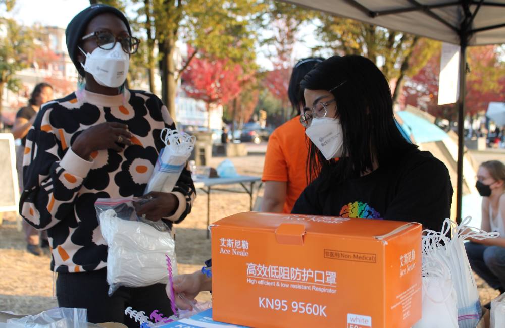 Two people are standing together against a stand of trees with orange leaves. The person to the left is wearing glasses, a floral sweater and a blue medical mask. The person to the right has black hair, glasses, and is wearing a black shirt with a white medical mask.