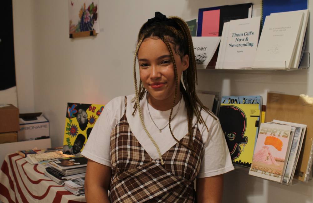 A woman with braided hair stands in front of a bookshelf at the Vancouver Black Library. She is wearing a white T-shirt under a light yellow and black Gingham dress. She is smiling at the camera.