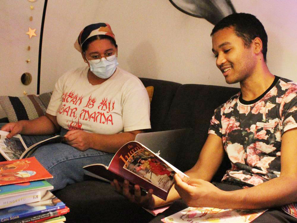Two young adults are seated on a dark sofa with stacks of books on the coffee table before them. The person on the left is wearing glasses, a medical mask and a white T-shirt with red text and jeans. The person on the right is wearing a black T-shirt with a pink floral pattern. They are holding up a book and smiling at its pages while their companion looks towards their book.