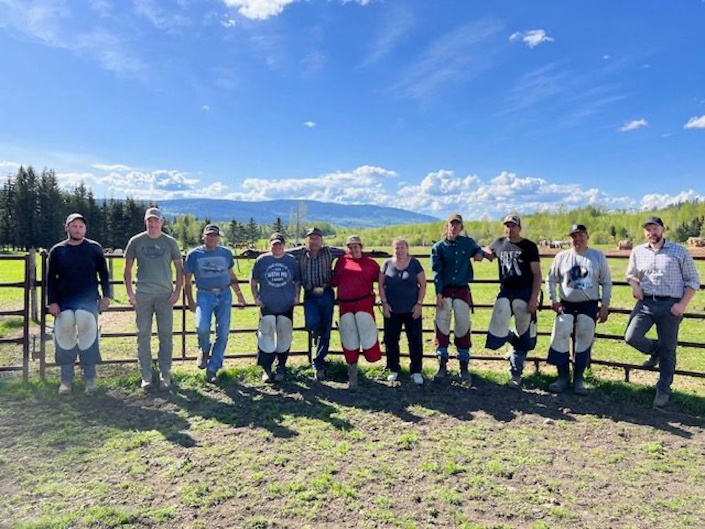 Eleven people line up along a fence for a photo on the ranch, on a sunny day.