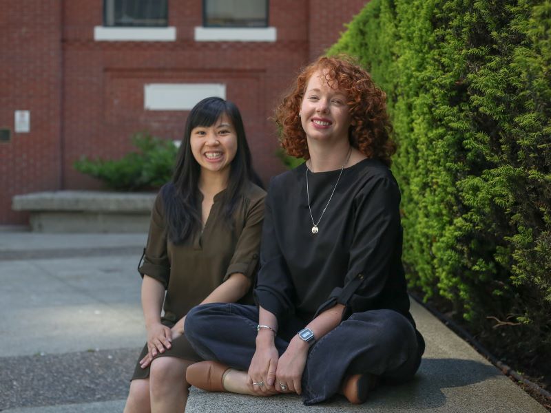 Kaitlyn Fung, left, and Kate Helmore, right, sit on a low concrete wall beside an evergreen hedge.