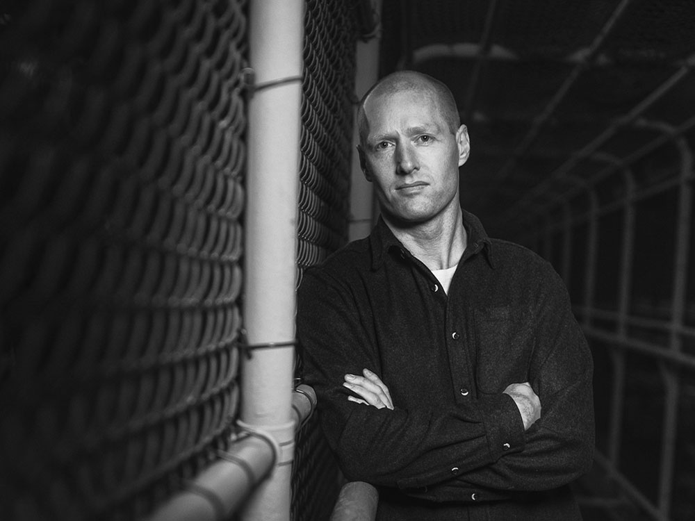 A black-and-white portrait of Vancouver-based journalist Arno Kopecky. He leans against a chainlink fence looking unsmilingly at the camera. He has a shaved head and a black shirt buttoned over a white T-shirt.