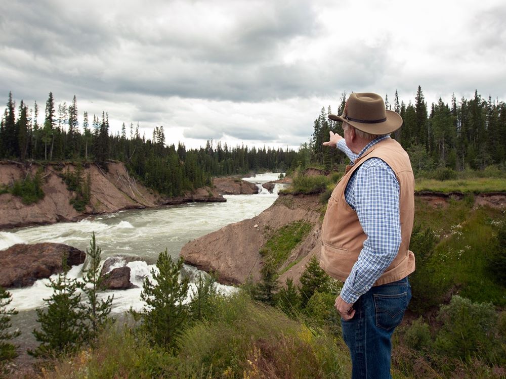 A lighter skinned older man with grey hair stands on the banks of a fast rushing river pointing at a short water fall up ahead and to his right. He is wearing a tan-coloured canvas vest over a blue striped shirt and a brown cowboy hat. The winding river is hemmed in by evergreen trees as well as tall grasses and shrubs.