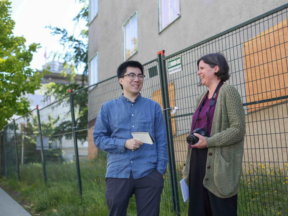 Two people are standing in front of a dark construction fence in front of a grey apartment building with its first-floor windows boarded up with plywood. Christopher Cheung, left, has glasses, black hair and a blue button-down shirt. Jen St. Denis, right, has short brown hair, a purple top and a long green cardigan. She has a camera around her neck and both are holding notebooks. The building stands in tall green grass. Trees and a residential downtown streetscape are in the background.