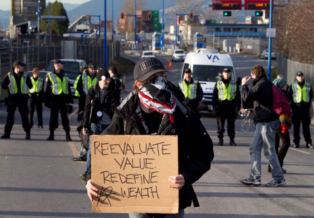 A protester wearing a black baseball cap and a bandanna over their face stands in the street holding a cardboard sign that reads 'Re-evaluate value. Redefine wealth.' In the background is a line of officers in neon green vests over black uniforms.