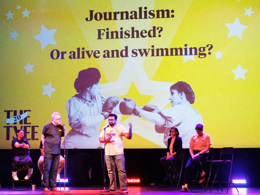 Steve Burgess, a man with light skin tone and silver-grey hair wearing a black T-shirt, and Mo Amir, a man with medium-dark skin tone, black hair and beard and glasses wearing a yellow T-shirt, stand onstage with microphones in front of them. Four other debaters are seated behind them. A large screen backdrop says "Journalism: Finished? Or alive and swimming?" in black type on a yellow background dotted with white stars.