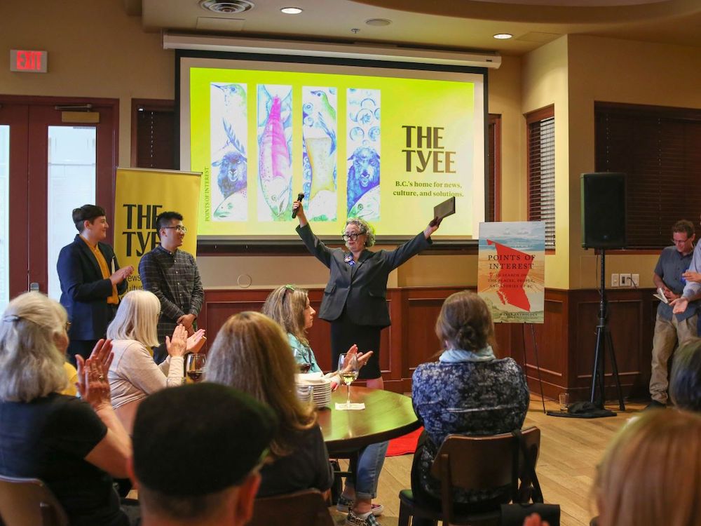 The audience looks on at Steamworks in Vancouver as Dorothy Woodend finishes her reading and throws her arms into the air. To her right are Christopher Cheung and andrea bennett. To her left is J.B. MacKinnon.