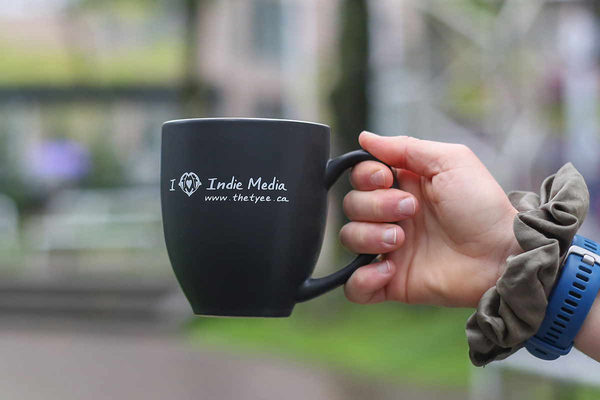 A person holds a black mug with 'I heart Indie Media' and 'www.thetyee.ca' in white lettering.