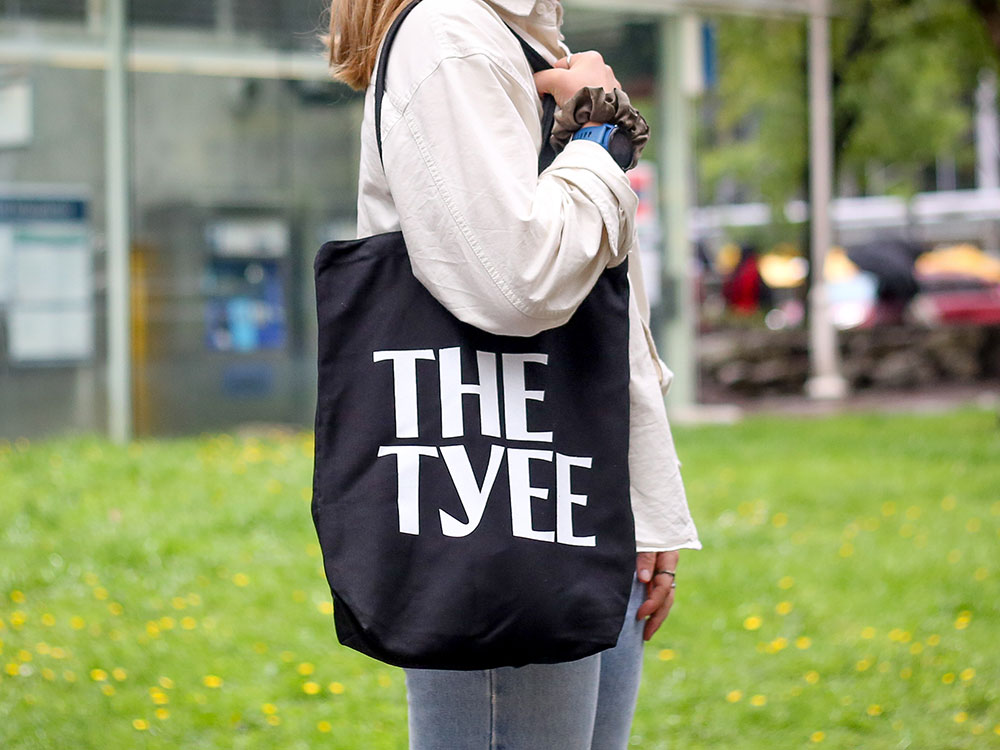 A close-up of a person’s torso standing in a park with a black tote that reads 'The Tyee' on their shoulder.