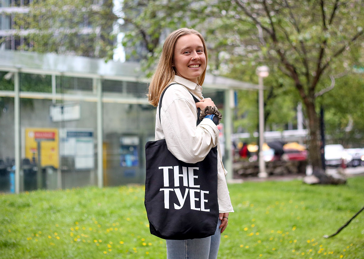 A woman with light skin, blond hair and a nose ring stands on the grass with a black tote bag over her shoulder that reads 'THE TYEE' in white all-caps letters.