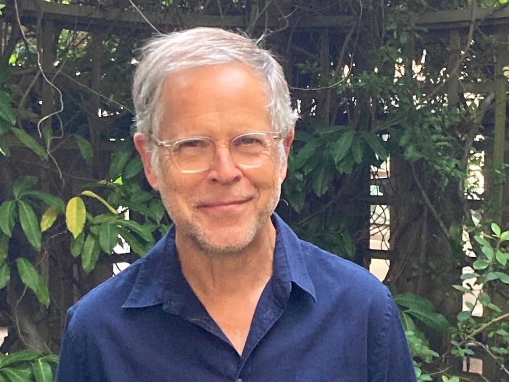 A middle-aged man with grey-white hair, light skin tone and clear glasses smiles at the camera. He is wearing a blue button-up shirt with the top button undone.