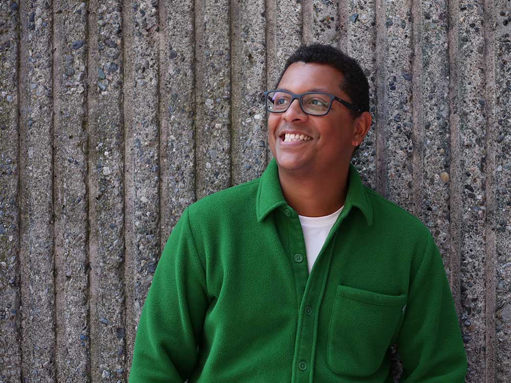 A man with medium-dark skin stands against a concrete wall in a forest green button-up fleece. He is wearing glasses, smiling broadly and looking upwards.