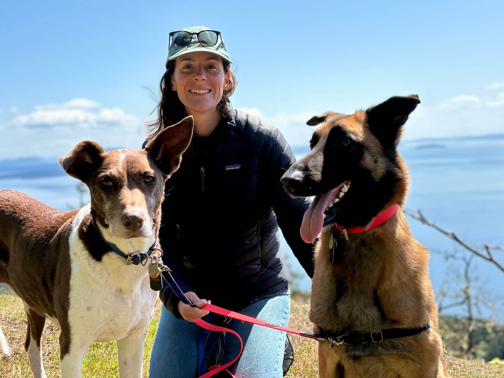 Carla Pellegrini, a woman with light skin tone and long brown hair, wears a ball cap and a black Patagonia coat and poses by the seaside with two dogs.