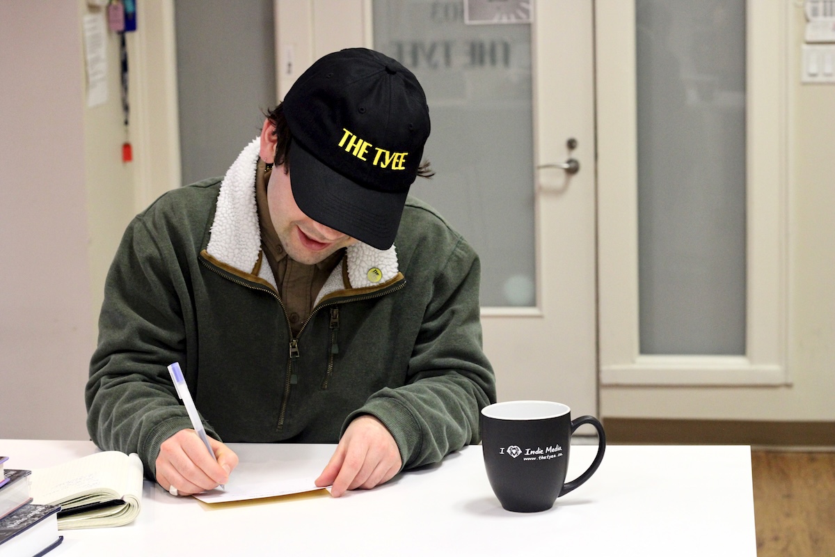 A reporter wearing a black &lsquo;The Tyee&rsquo; baseball cap takes notes with pen and paper.