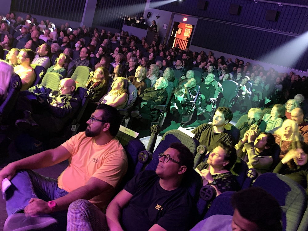 A full theatre with audience members lit up by purple, yellow and green lights.