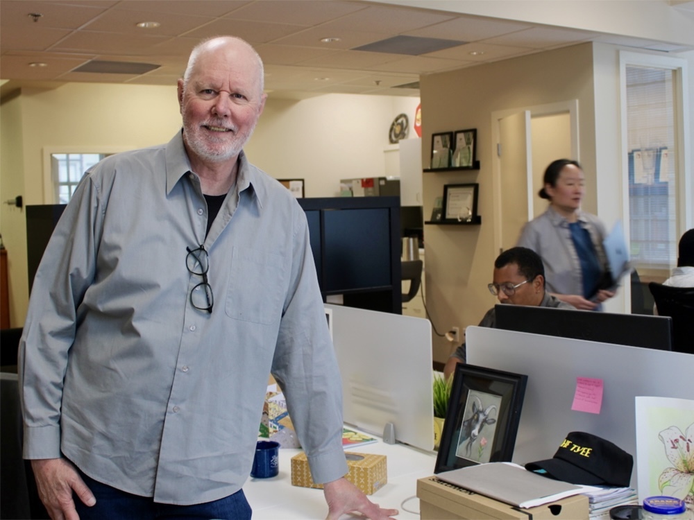 In the foreground, David Beers, a late middle-aged man, balding with a grey beard and wearing a grey open collar shirt with blue jeans, smiles at the camera. In the background, Tyee staffers are visible doing their work.