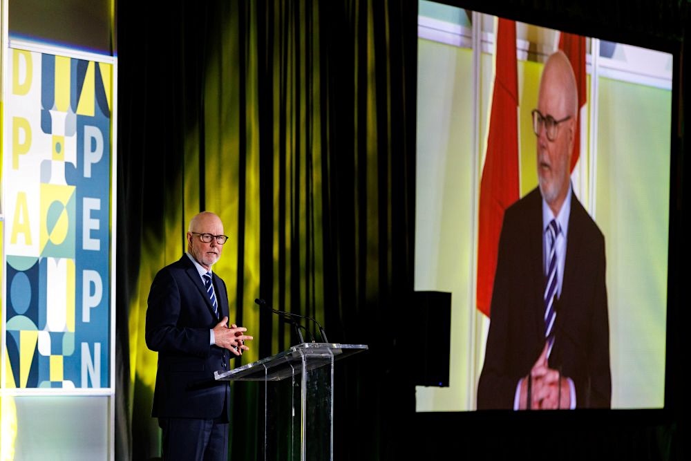 A man in his mid-60s wearing a suit stands at a podium. Behind, a screen shows a large video image of him.