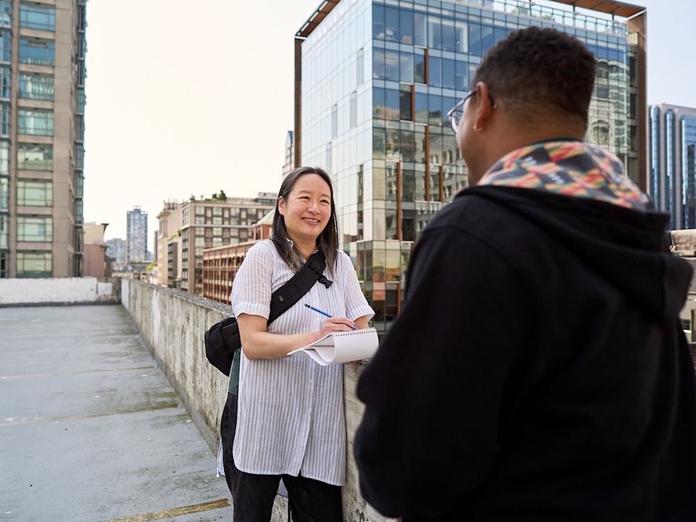 A woman with long black hair wears a white and blue striped blouse and smiles at a man who stands with his back to the camera. The woman is holding a notebook and a pen. They are outdoors with office buildings around them.