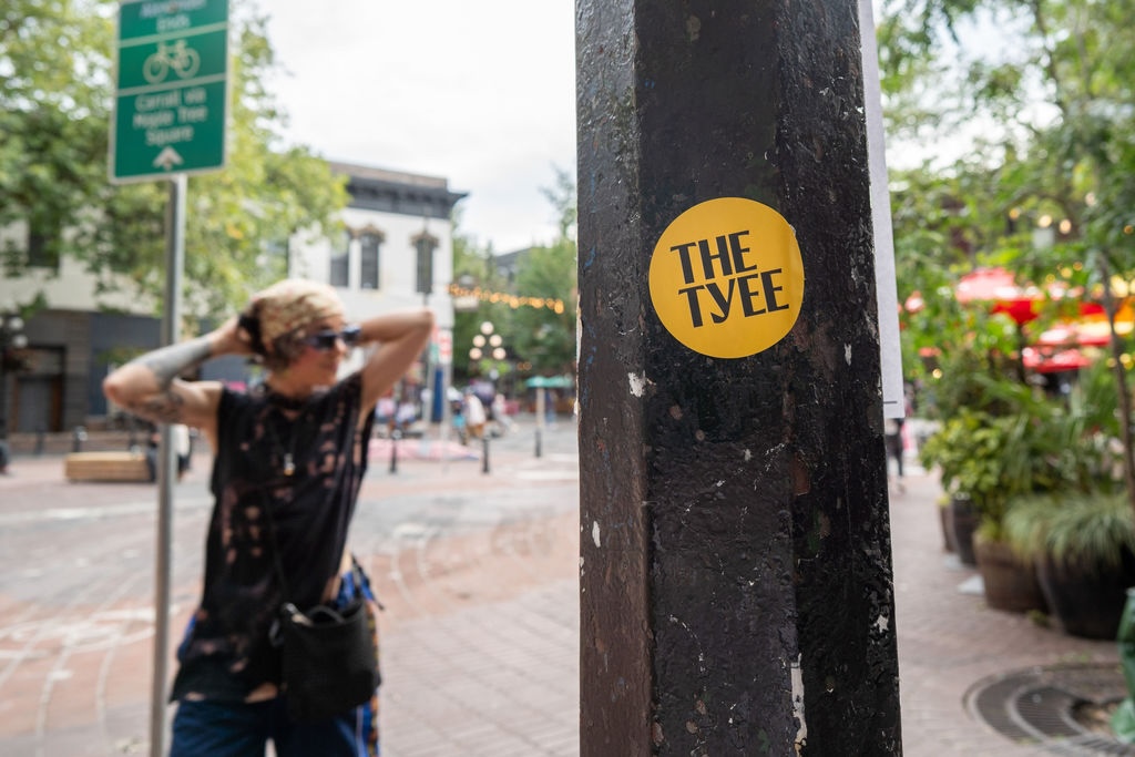 A yellow circular sticker is stuck to a black pole in a city street. A person in casual streetwear stands in the background.