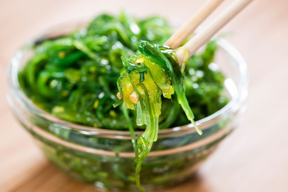 Close-up of chopsticks holding strings of bright green seaweed salad, with a bowl of the salad out of focus in the back.
