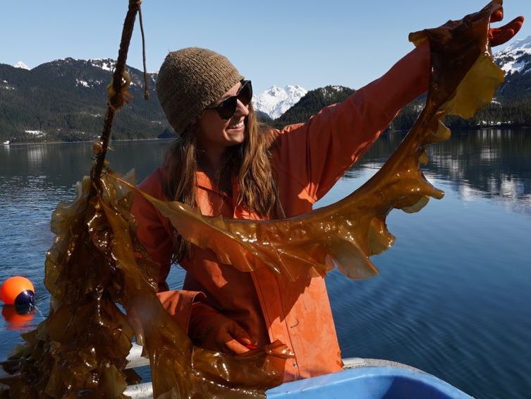 A woman with light skin and dark reddish hair, wearing dark sunglasses, a brown toque and an orange slicker, holds up a string of brown kelp. Water and snowy mountains can be seen behind her.