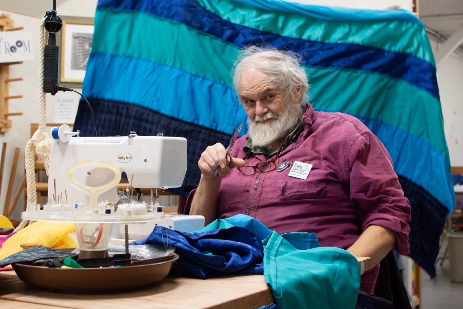 Harold Niven sits to the left of the frame and looks wryly at the camera, a pair of sewing scissors in hand. He has light skin, unruly white hair and a white beard. A red pair of glasses sits around his chest against a burgundy button-down shirt. He sits at a sewing table before a white sewing machine and a swath of turquoise and blue fabric. Behind him is a large, striped, blue, turquoise and dark blue blanket hanging on a wide high structure.
