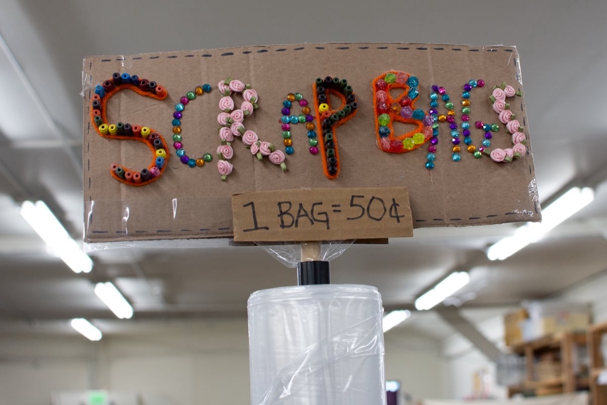 In an interior space with white fluorescent lights on the ceiling, a handmade cardboard sign that reads 'Scrap bins, 1 bag = 50 cents' stands atop a roll of clear plastic bags. The phrase 'Scrap bins' is decorated with a colourful, bric-a-brac assortment of beads, gem stickers and small pink fabric roses.
