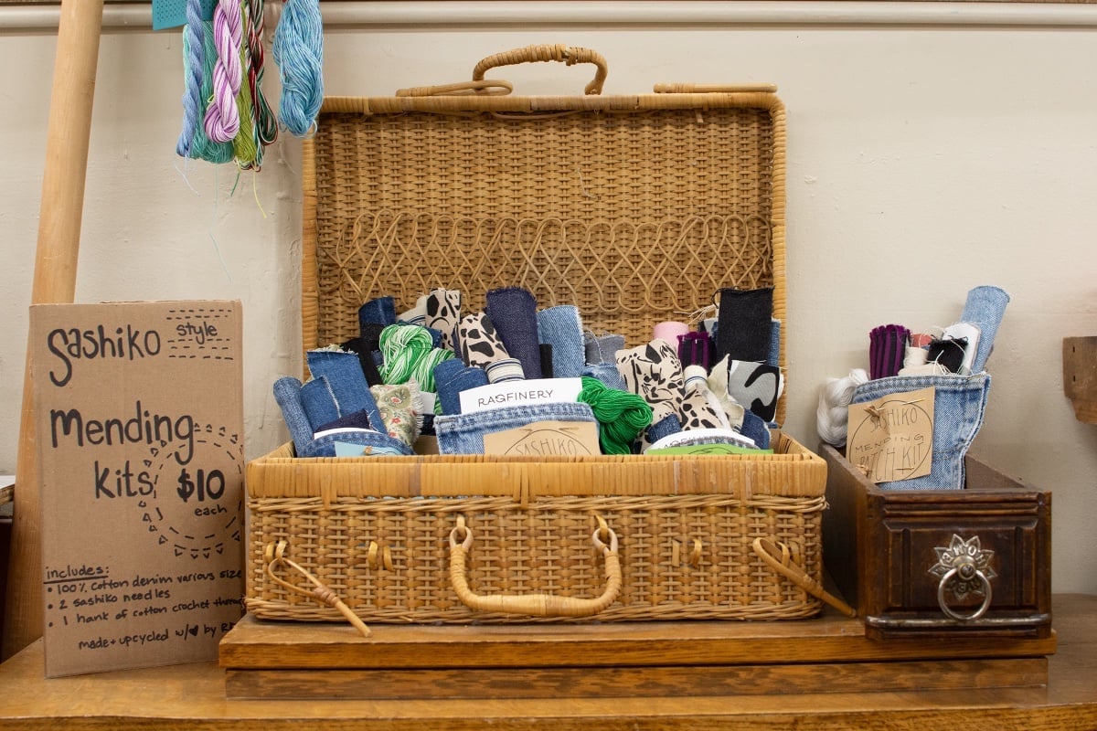 A small wicker basket sits at the centre of the frame atop two small wood boards. To its right is a rectangular wooden box with a silver fixture on its front. Both the box and the basket are standing open to reveal several small bundles of rolled-up fabric in upcycled denim pouches. A handmade cardboard sign to the left explains that they are sashiko mending kits.