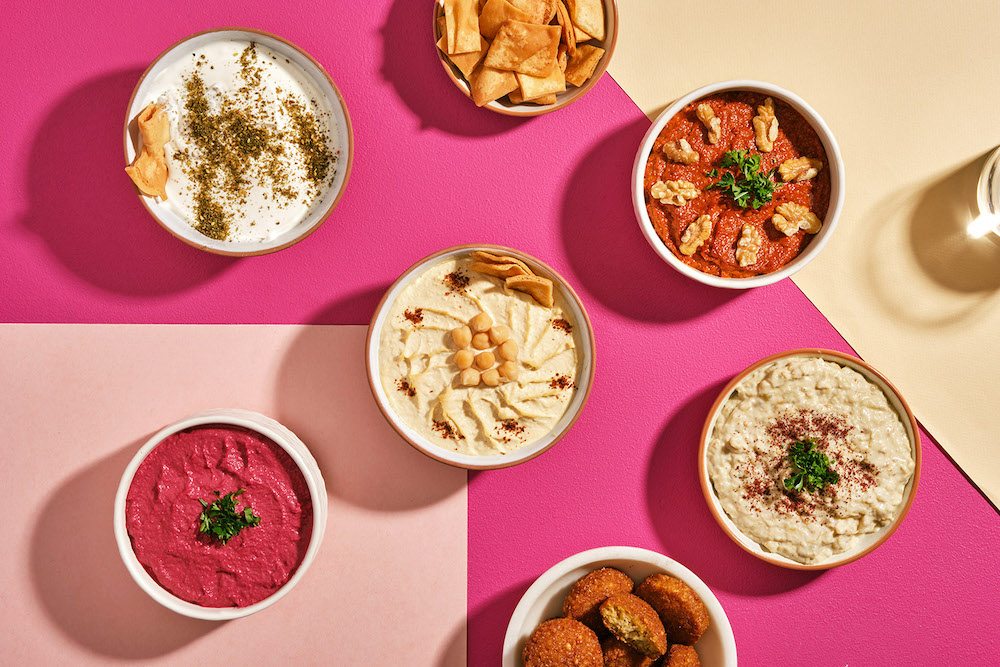 Bowls of dips, pita chips and falafel are arranged across a colourful table with checkerboard fuchsia colourways. The dishes are photographed from above.