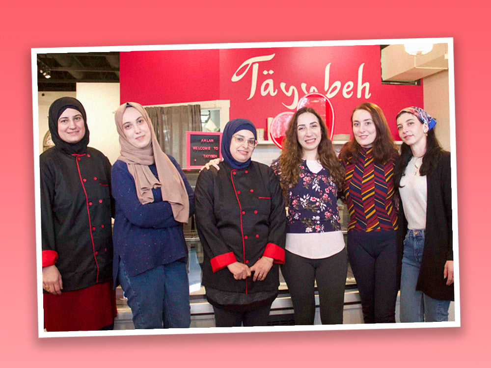 Six light-skinned women stand together in the front section of a newly opened restaurant against a fuchsia wall with 'Tayybeh' written on it. They are wearing casual streetwear or black chef’s outfits, smiling at the camera.