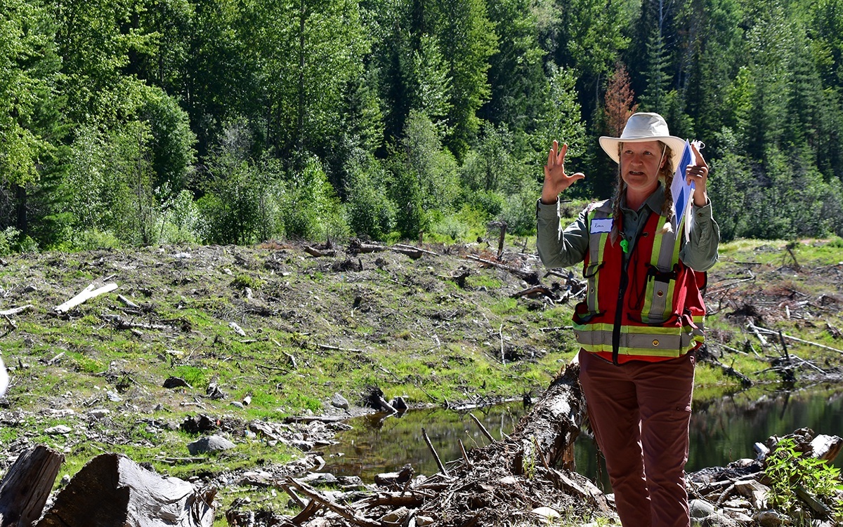 A middle-aged white woman wearing a broad-brimmed tan hat and a high-visibility vest gestures. Behind her is a marshy area.