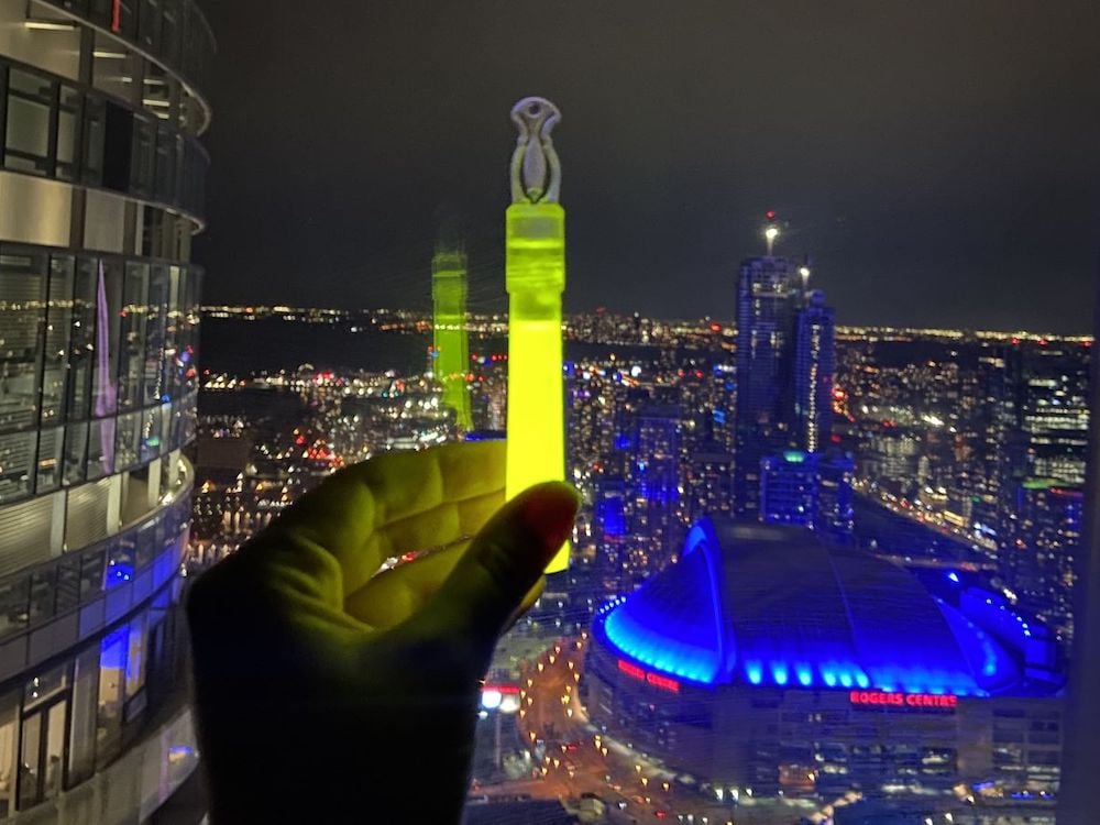 A hand holds up a glowing translucent stick in front of a window at night. Outside is a lit city skyline.