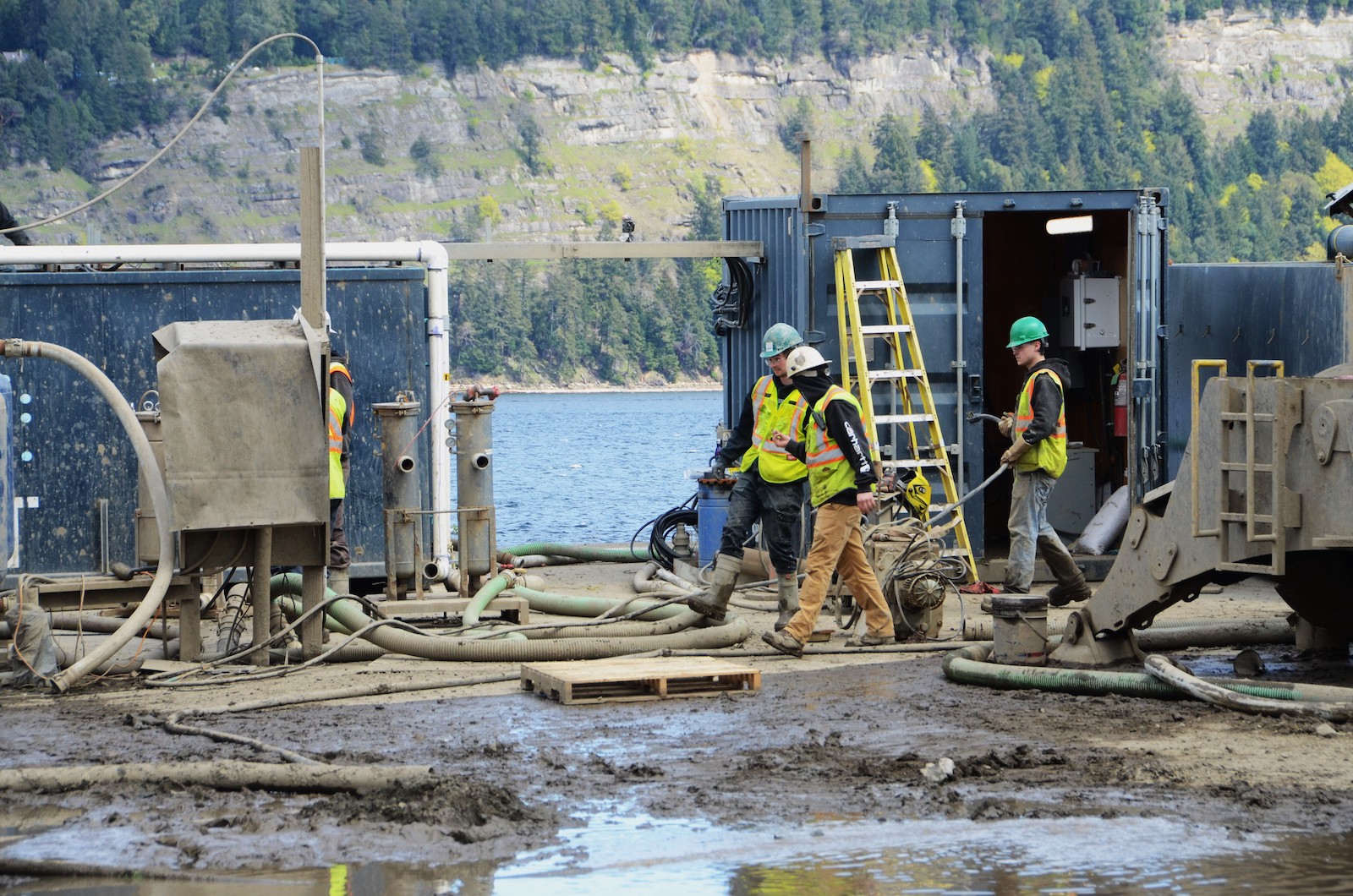 Workers in hard hats and high-vis vests walk through the muddy yard of a plant on the water’s edge.