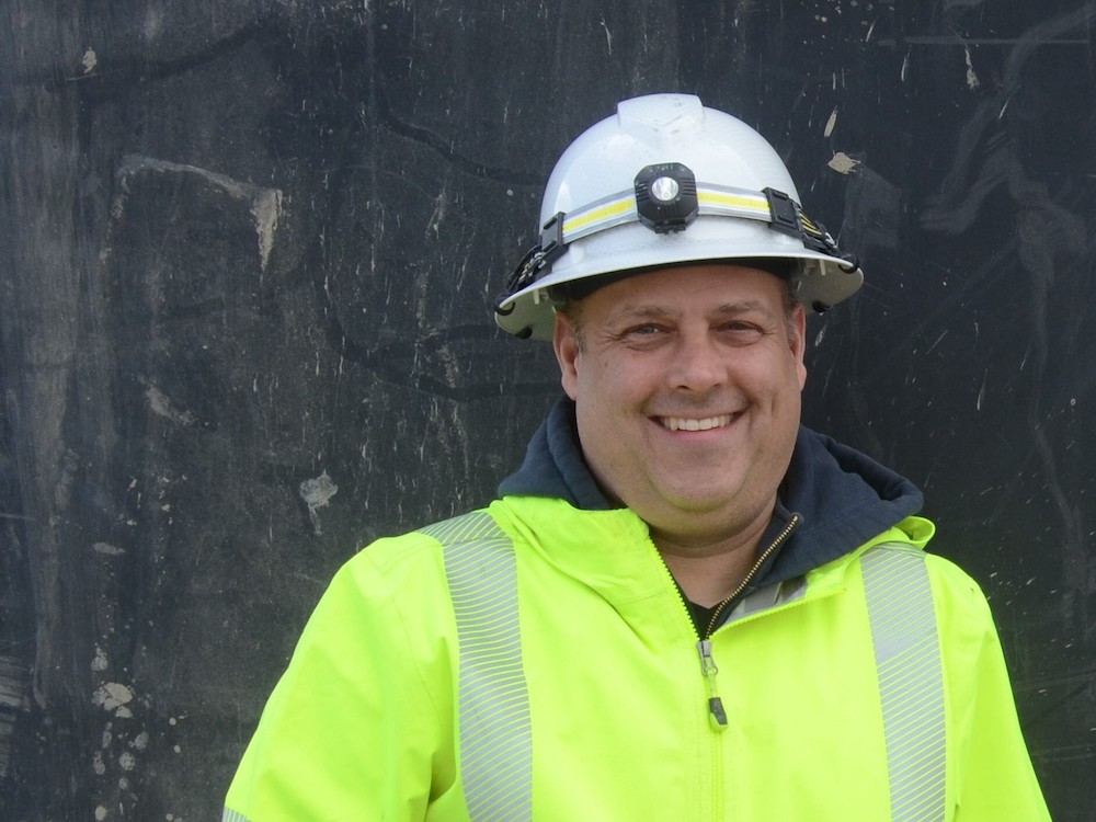 A light-skinned man wearing a white hard hat and high-vis jacket smiles into the camera.