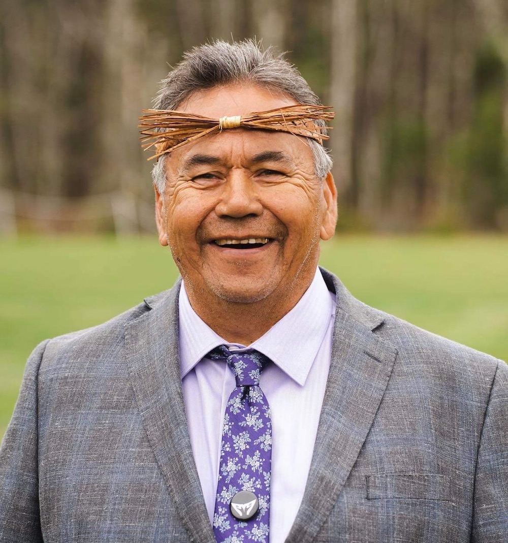 A man with medium skin tone wears a small traditional headband made from wood, a purple tie and a grey suit jacket.