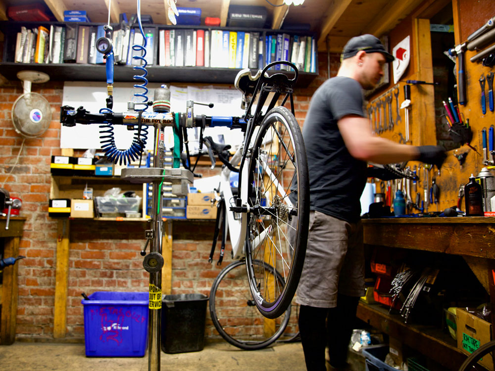 A bicycle is suspended on a bike stand in a small room with bike tools, parts and manuals on the walls, side tables and floor. To the right of the bike stands a man with medium-light skin sorting through objects on a table.