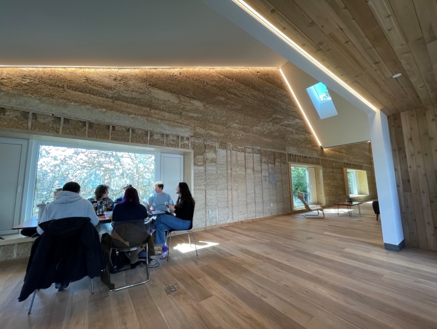 A group of students sit at a table in a spacious room with wood floors and hempcrete walls.