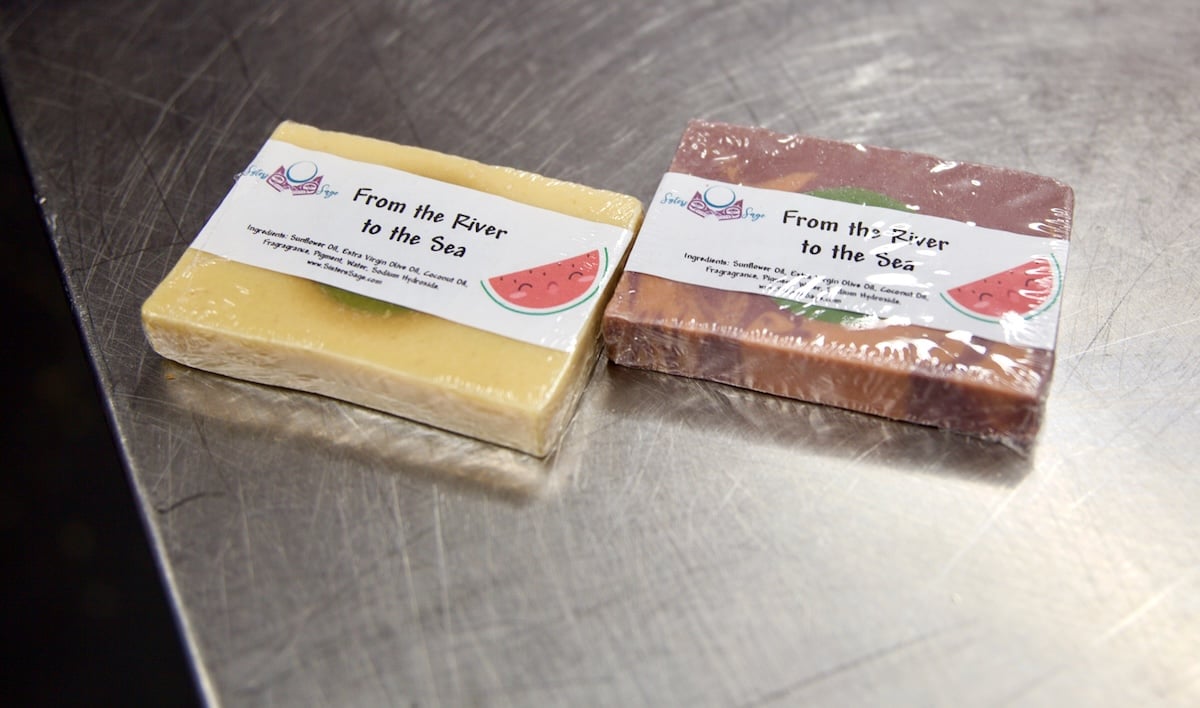 Two bars of individually wrapped squares of soap lie on a stainless steel tabletop. The soap labels read ‘From the River to the Sea.’