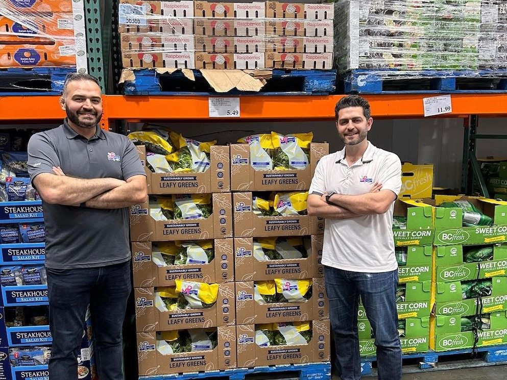Two men with medium-light skin tone, dark hair and beards stand with arms akimbo amid stacks of food boxes on pallets.