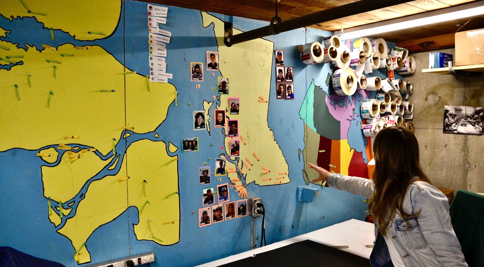 Colourful maps of the Fraser River delta, B.C. and Canada are painted onto a wall. Nearly two dozen photos of fishers are tacked on around the map of B.C.
