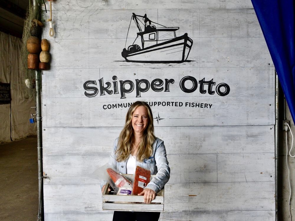 A smiling woman with light skin tone and long blond hair holds a wooden crate full of packaged seafood. Behind her is a sign that says ‘Skipper Otto Community-Supported Fishery’ with an illustration of a small fishing boat.