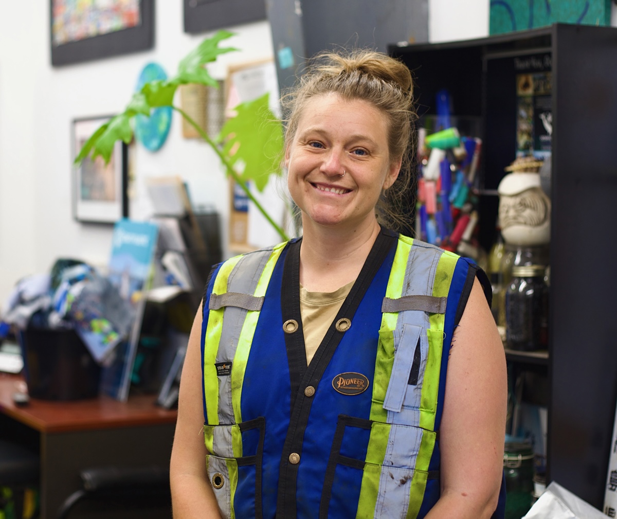A woman with light skin tone and reddish-blond hair pulled into a ponytail smiles at the camera. She is wearing a blue and yellow reflective safety vest and a light brown T-shirt.