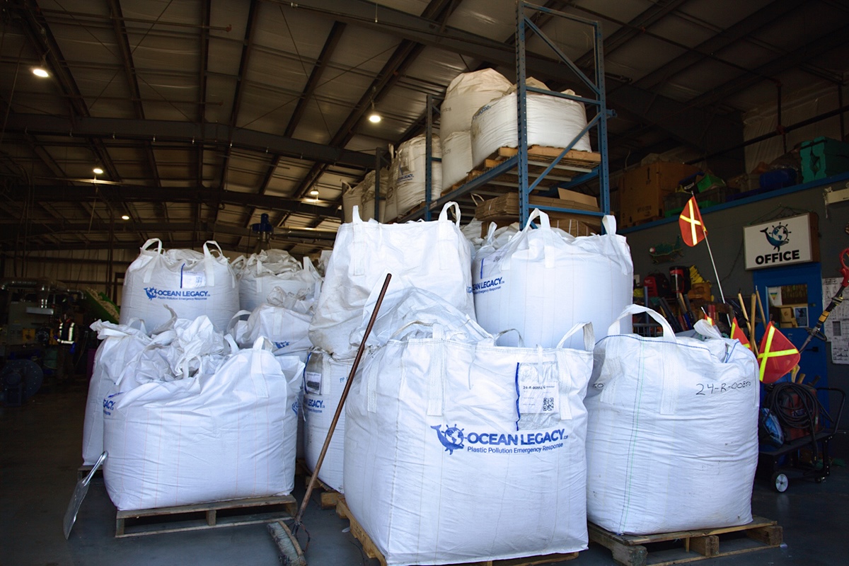 Dozens of large, cube-shaped plastic sacks full of processed plastic recyclables are stacked on a concrete floor and metal shelves.