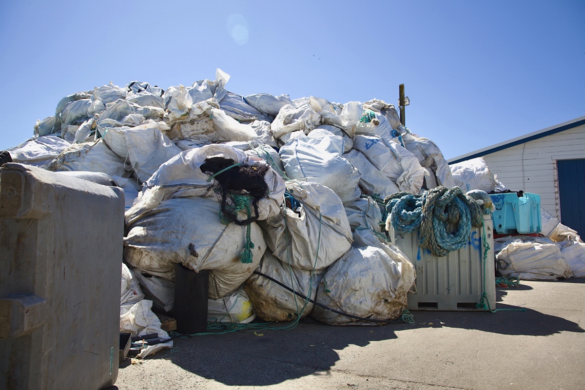A pile of white garbage bags filled with ocean-recovered rope and other plastics, almost as tall as a nearby white shed. A grey fish tote lying on its side is in the foreground.